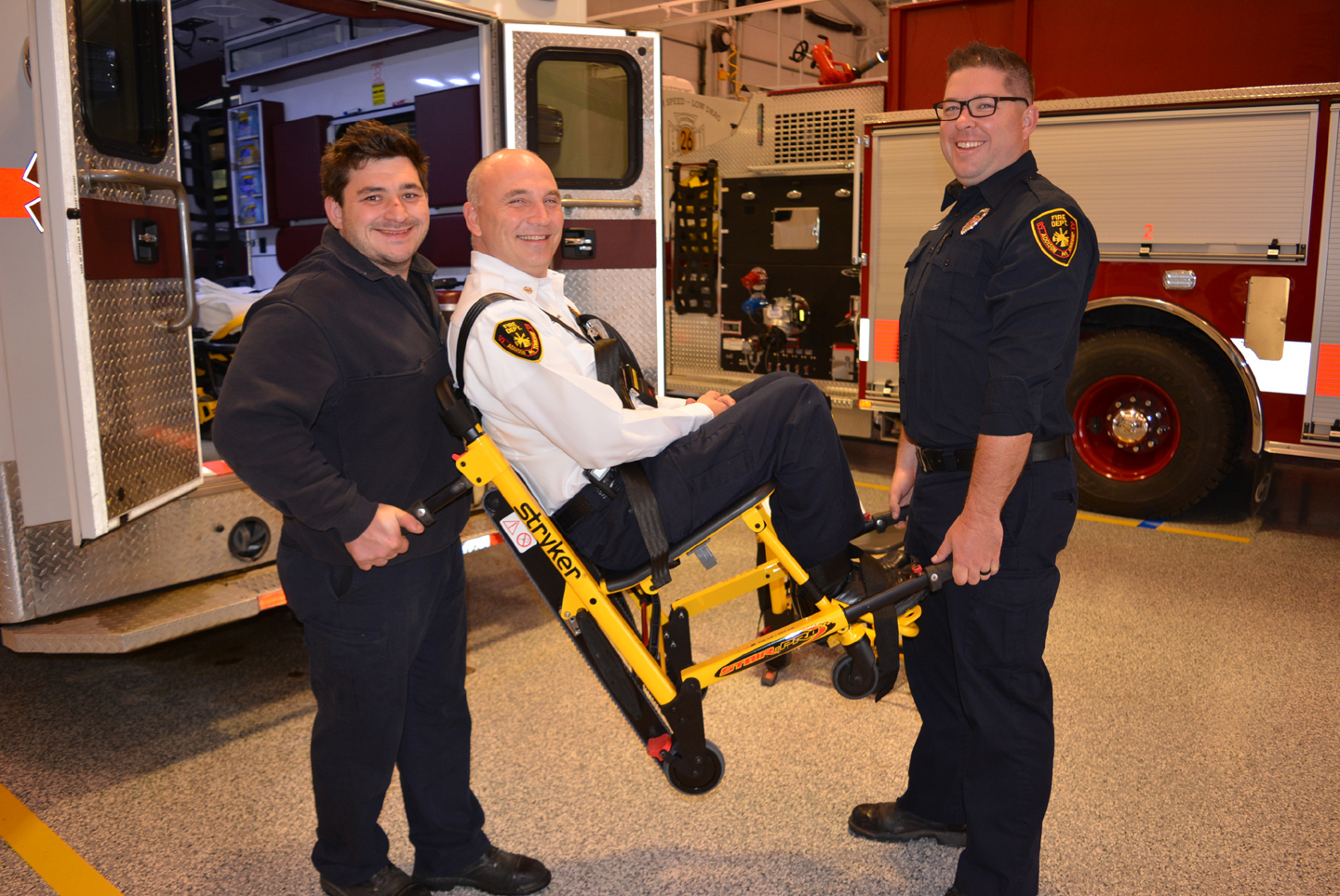 Addison Township Firefighter Tyler Hunsucker (left) and EMS Coordinator Rob Fitzpatrick (right) demonstrate how easy it is to lift someone, like Fire Chief Jerry Morawski, with the department’s new stair chairs. Photo by C.J. Carnacchio. 